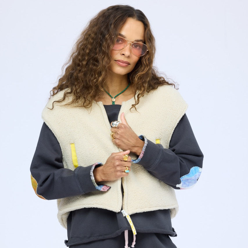 Woman wearing a cream sherpa vest with colorful patchwork sleeves over a navy studio sweatshirt, with sunglasses and a necklace.