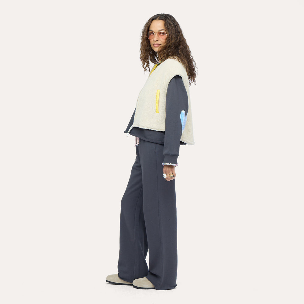 Woman wearing a cream sherpa vest with colorful patchwork sleeves over a navy sweatshirt and navy sweatpants, with sunglasses.