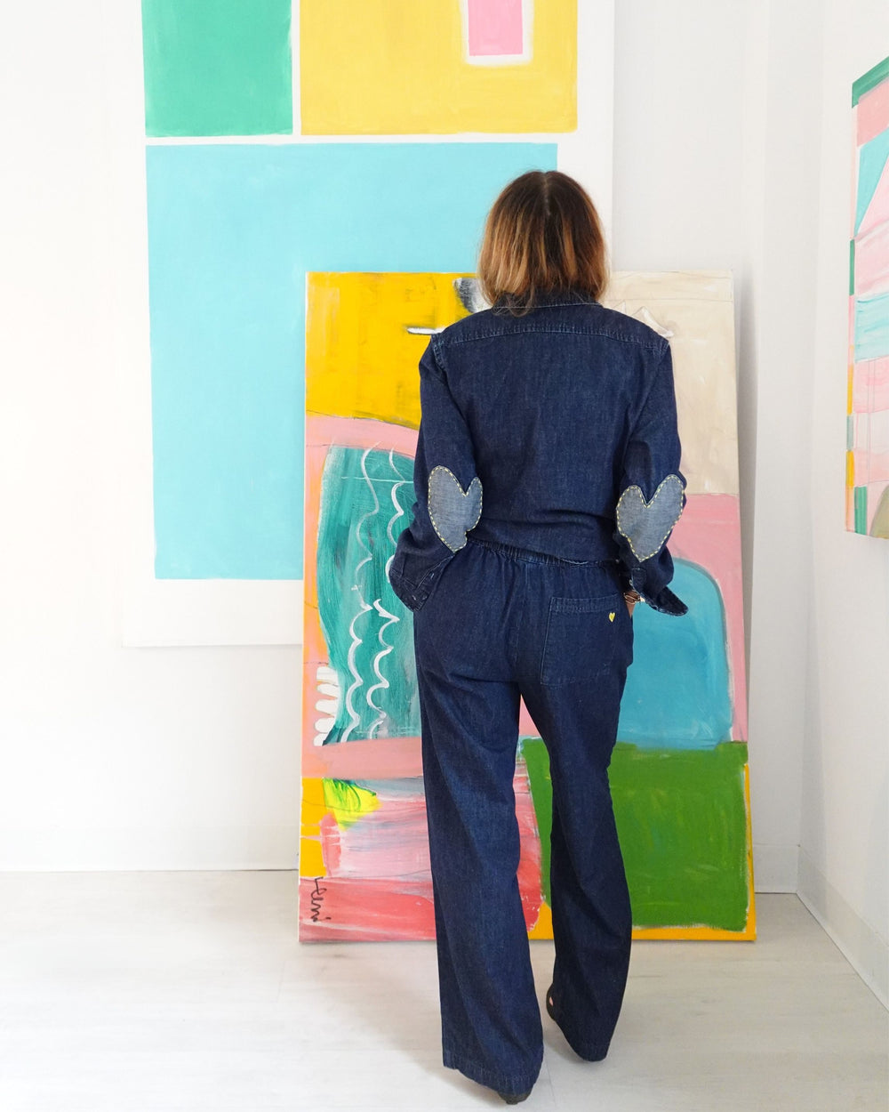 Woman wearing a dark indigo denim  wide-leg palazzo pants, a collared button-up top with heart-shaped elbow patches, and an elastic waist with patch pockets, seen from behind against colorful abstract paintings