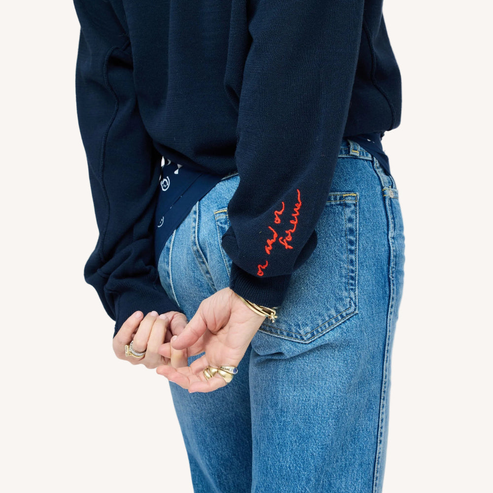 Close-up of a woman wearing a Kerri Rosenthal navy cardigan with red 'now and forever' embroidery on the sleeve, styled with blue jeans and a navy heart print bandana.