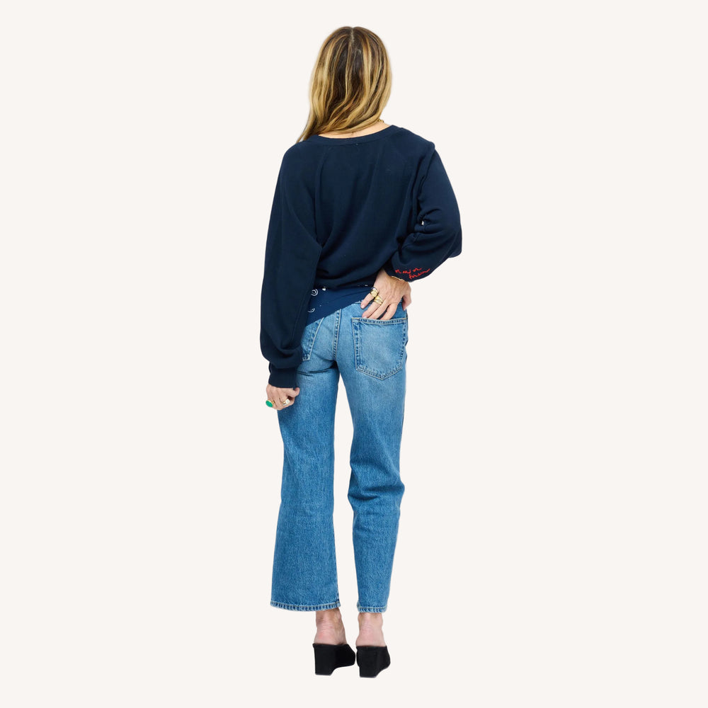Back view of a woman wearing a Kerri Rosenthal navy cardigan with red sleeve embroidery, styled with cropped blue jeans and black mules.