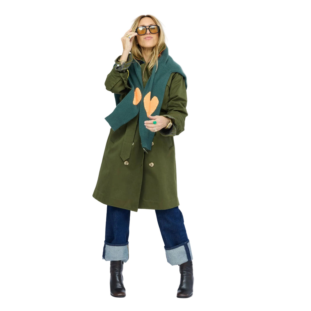 Woman layering the green cashmere sweater with orange heart elbow patches under an olive trench coat, styled with cuffed jeans and ankle boots
