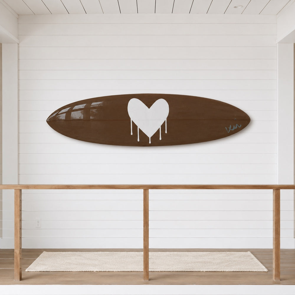 A brown surfboard-shaped decorative plaque or tray featuring a large white dripping heart in the center and a small white script signature on the right side, shown against a plain white background.