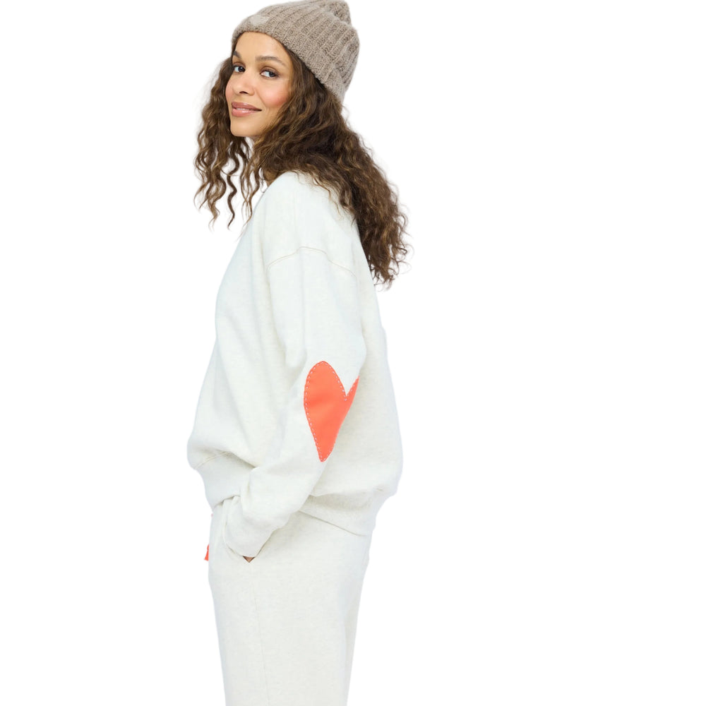 Woman in profile smiling over her shoulder, wearing an oatmeal heather sweatshirt with a coral heart elbow patch, matching sweatpants, and a taupe knit beanie.