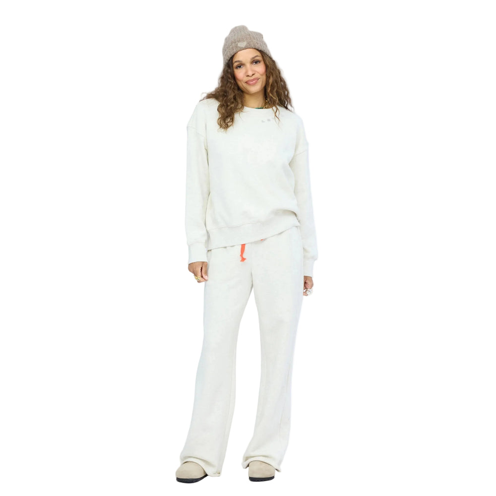 Woman facing forward wearing an oatmeal heather sweatshirt and matching wide-leg sweatpants with a coral drawstring, taupe slip-on shoes, a taupe knit beanie, and layered gold jewelry.
