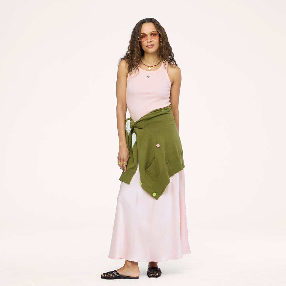 Model wearing a pale pink ribbed tank with a heart pendant necklace, an olive green cardigan tied around the waist, a blush satin maxi skirt, and black woven flats. Shop Kerri Rosenthal