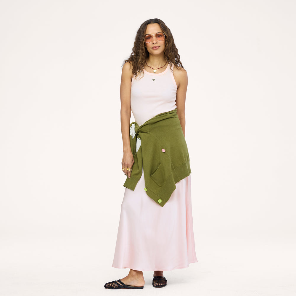 Model wearing a pale pink ribbed tank with a heart pendant necklace, an olive green cardigan tied around the waist, a blush satin maxi skirt, and black woven flats. Shop Kerri Rosenthal