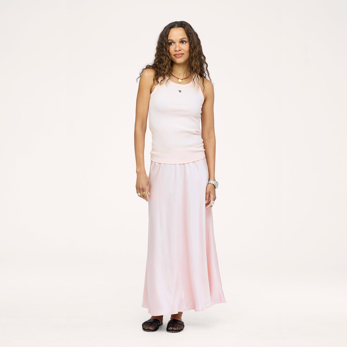Model wearing a pale pink ribbed tank with a heart pendant necklace and a blush pink satin maxi skirt, paired with black woven flats. Shop Kerri Rosenthal