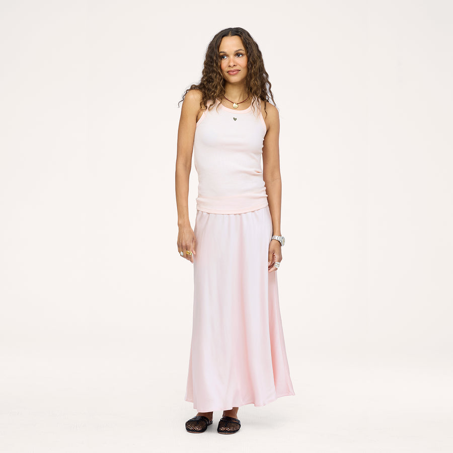 Model wearing a pale pink ribbed tank with a heart pendant necklace and a blush pink satin maxi skirt, paired with black woven flats. Shop Kerri Rosenthal