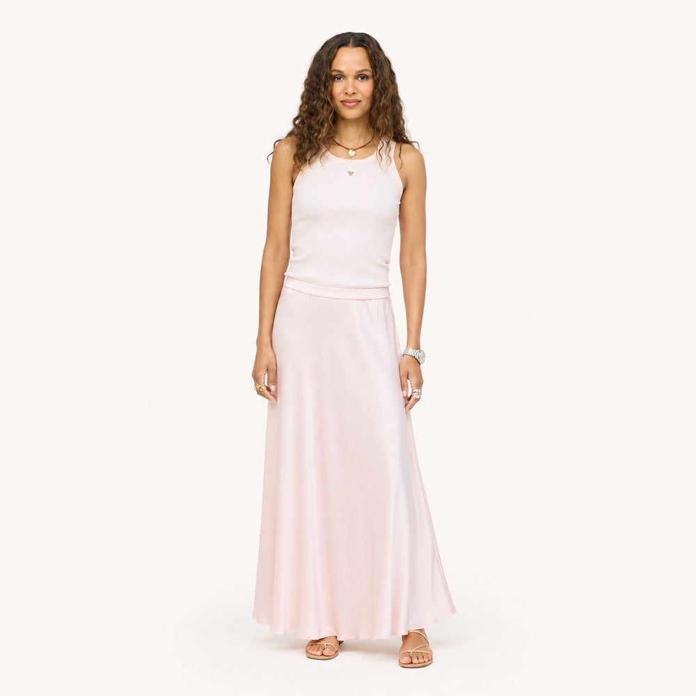 Model wearing a pale pink ribbed tank with a heart pendant necklace and a blush pink satin maxi skirt, paired with black woven flats. Shop Kerri Rosenthal