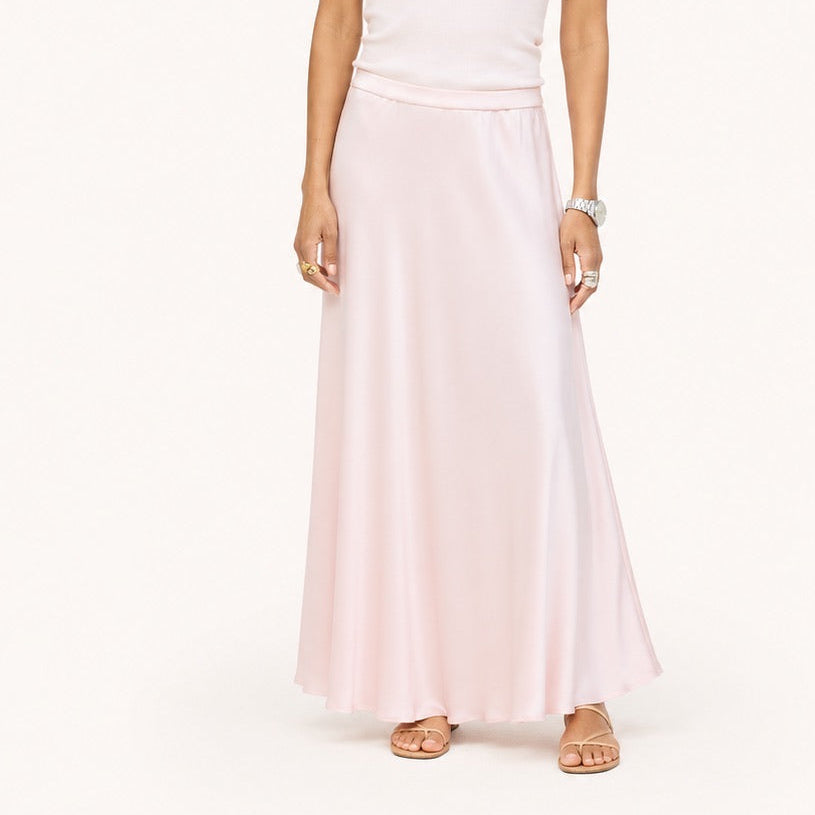 Model wearing a pale pink ribbed tank with a heart pendant necklace and a blush pink satin maxi skirt, paired with black woven flats. Shop Kerri Rosenthal