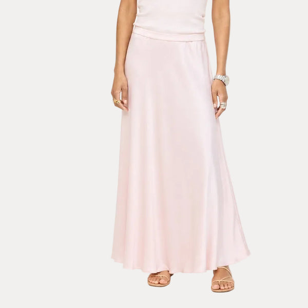 Model wearing a pale pink ribbed tank with a heart pendant necklace and a blush pink satin maxi skirt, paired with black woven flats. Shop Kerri Rosenthal