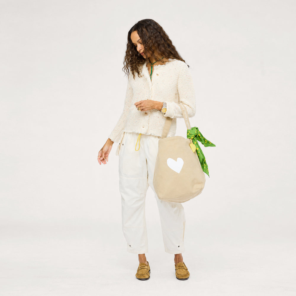 Woman in Kerri Rosenthal cream tweed jacket styled with white pants, tan loafers, and a KR heart tote bag with a green scarf.