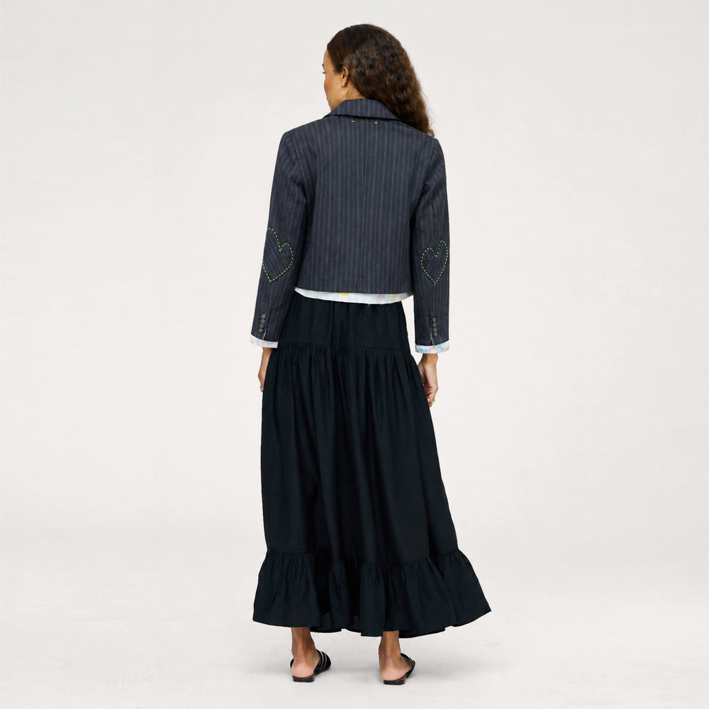 Back view of model wearing a navy pinstriped cropped blazer with heart-embroidered elbow patches over a black tiered maxi skirt