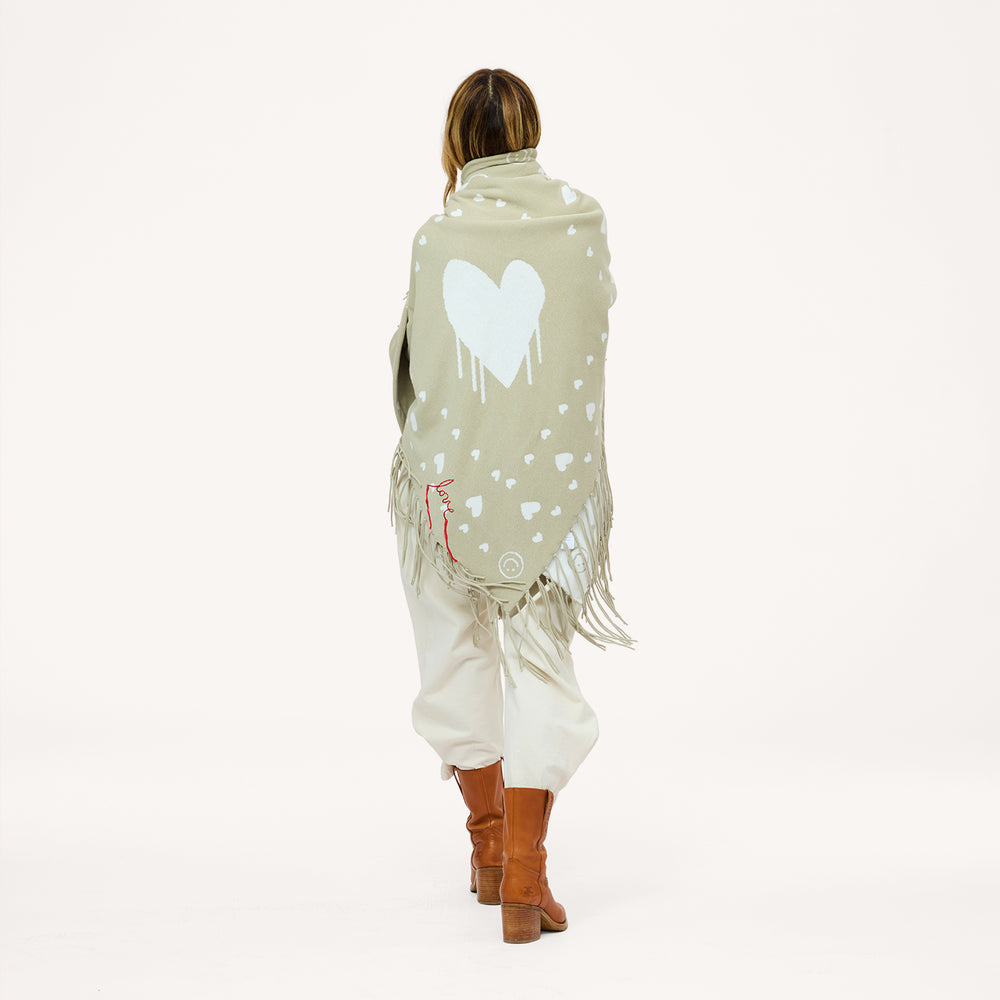 Back view of a woman holding up a sage green fringed poncho with a heart pattern and red 'love' stitching, wearing cream pants and brown boots.