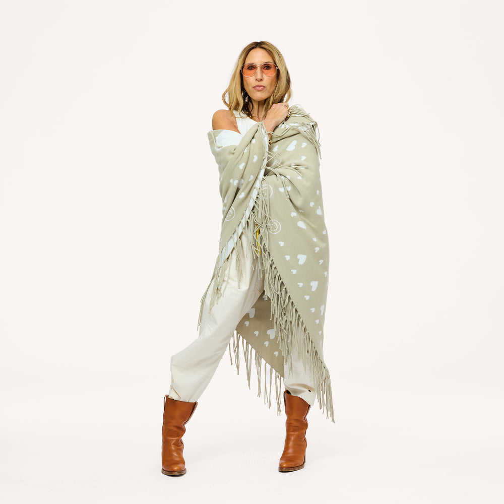 Woman wrapped in a sage green fringed poncho with a heart pattern, wearing cream pants, brown boots, and sunglasses.