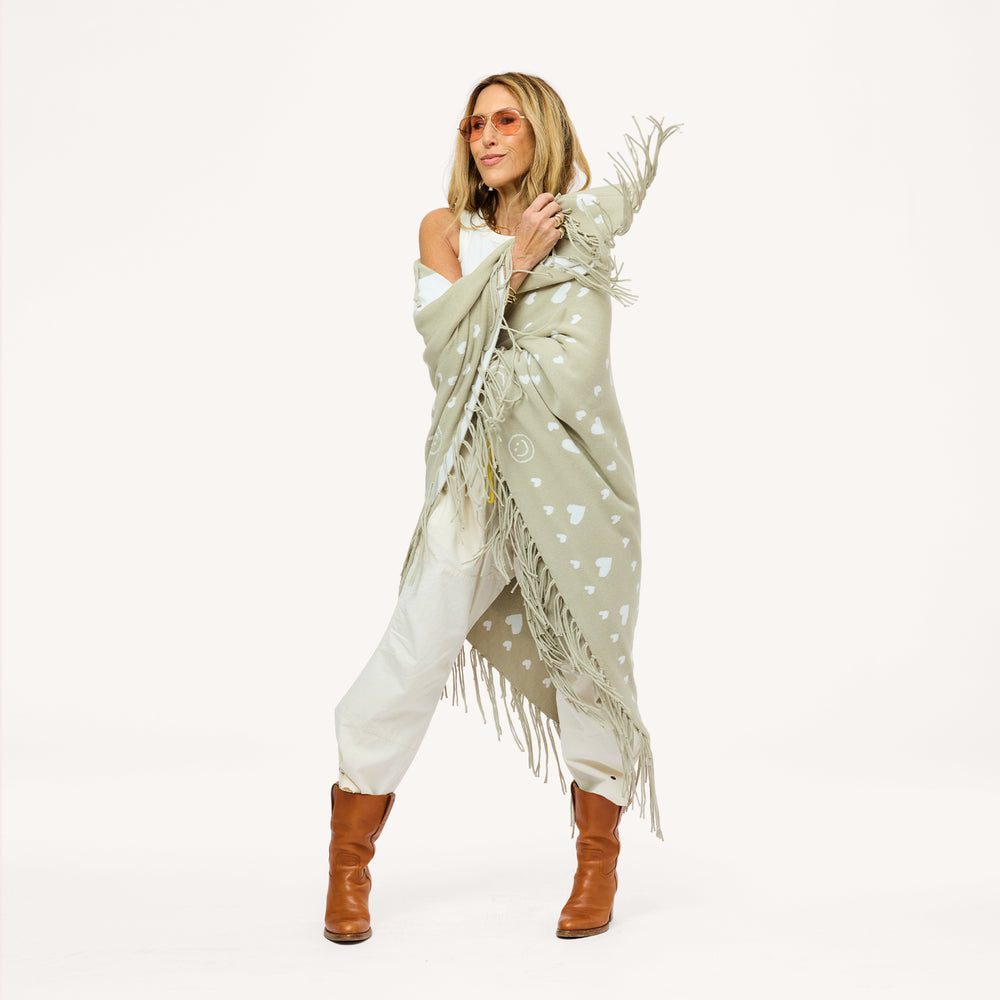 Woman wrapped in a sage green fringed poncho with a heart pattern, wearing cream pants, brown boots, and sunglasses.