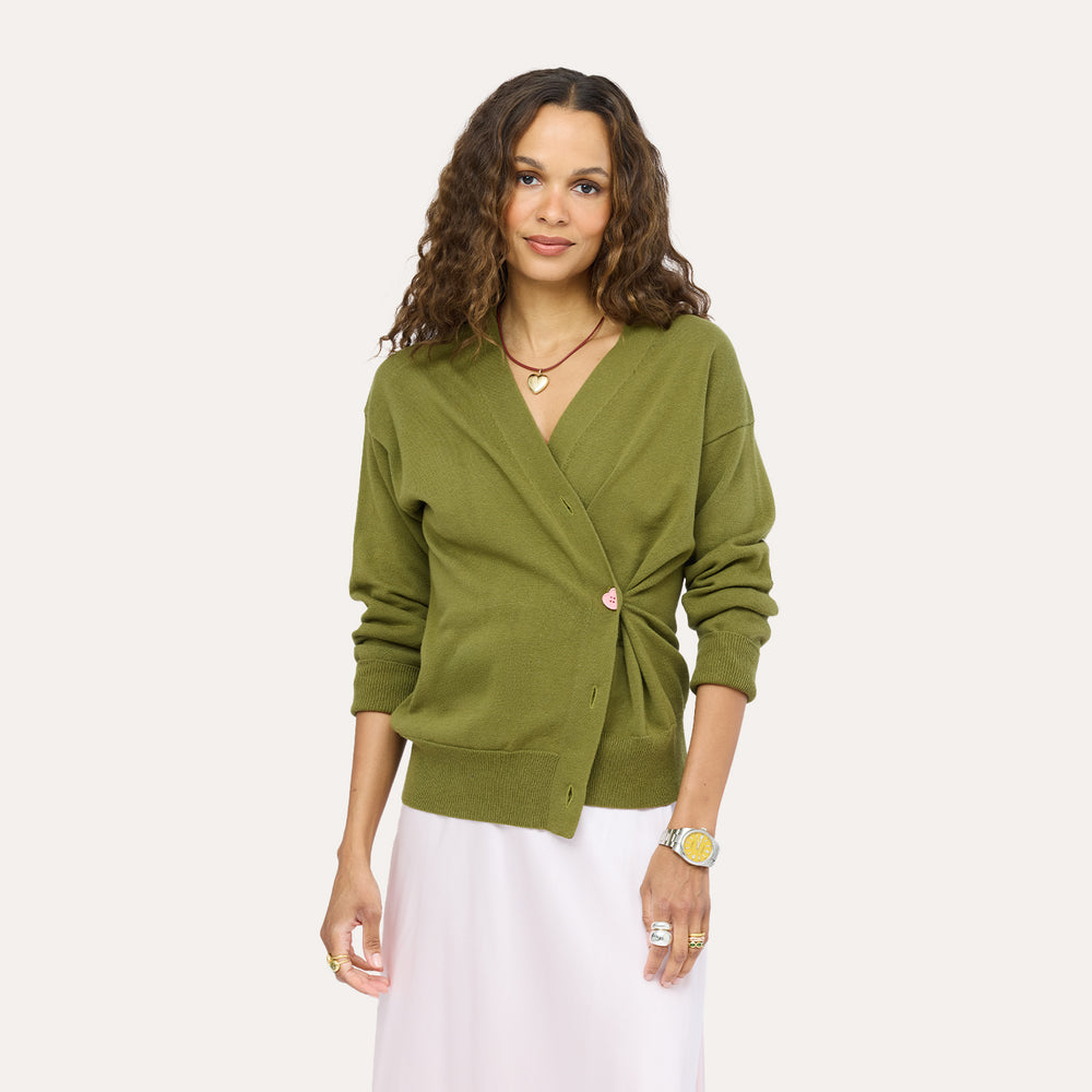 Close-up of a woman wearing a Kerri Rosenthal olive green wrap-style knit cardigan with a gold heart button, styled with a blush pink skirt and a heart pendant necklace.