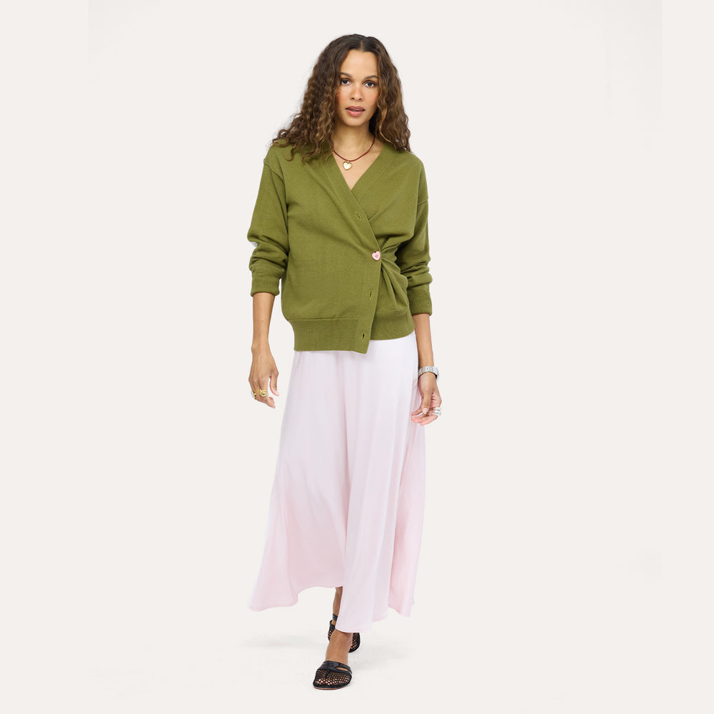 Woman wearing a Kerri Rosenthal olive green wrap-style knit cardigan with a gold heart button, styled with a blush pink maxi skirt and black woven loafers.