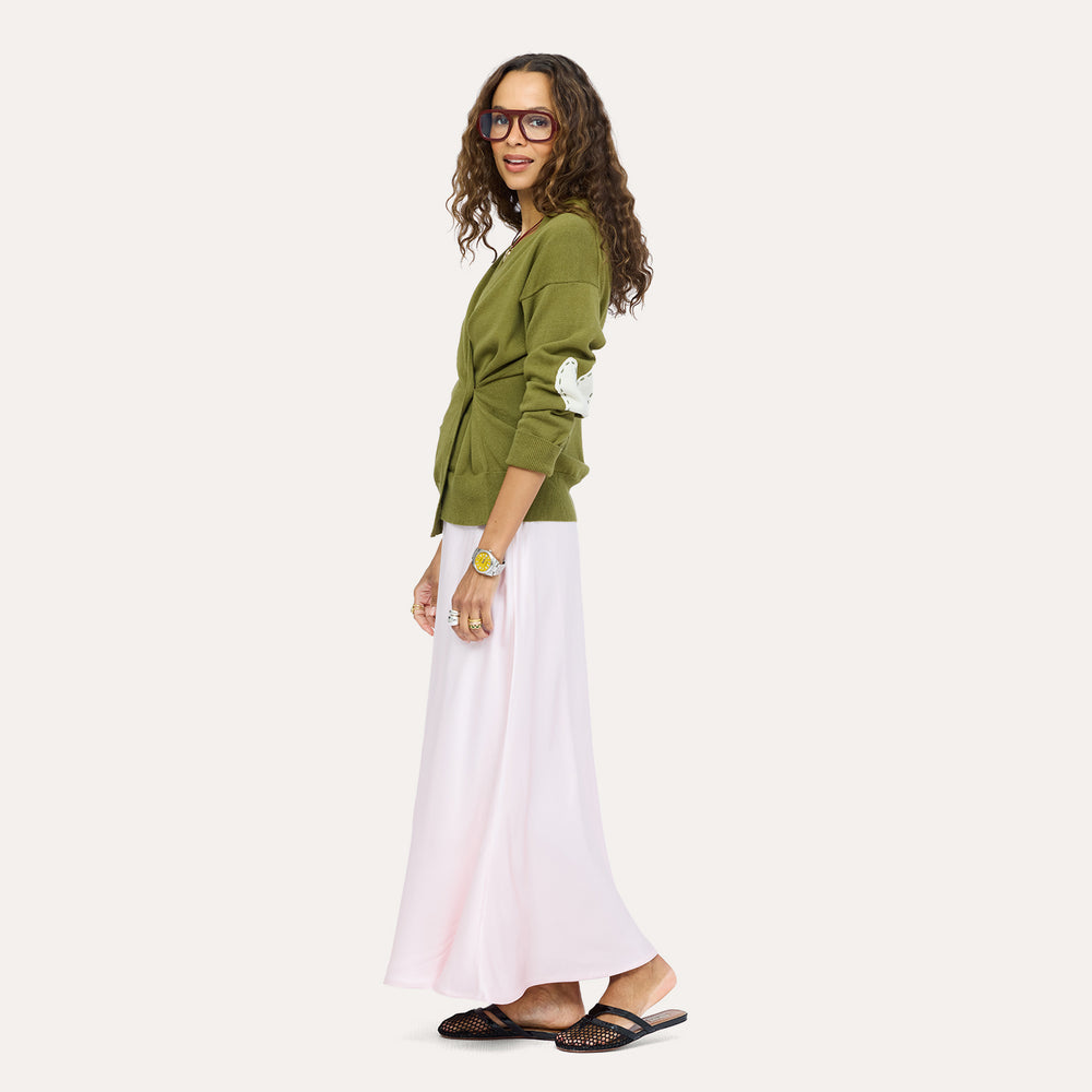 Side view of a woman wearing a Kerri Rosenthal olive green knit cardigan with mint green heart elbow patches, styled with a blush pink maxi skirt, square frame glasses, and black woven sandals.