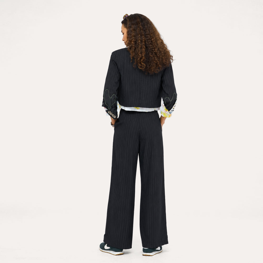 Back view of woman wearing a black pinstripe blazer and pants with paint splat cuffs, a pink top, and sneakers.