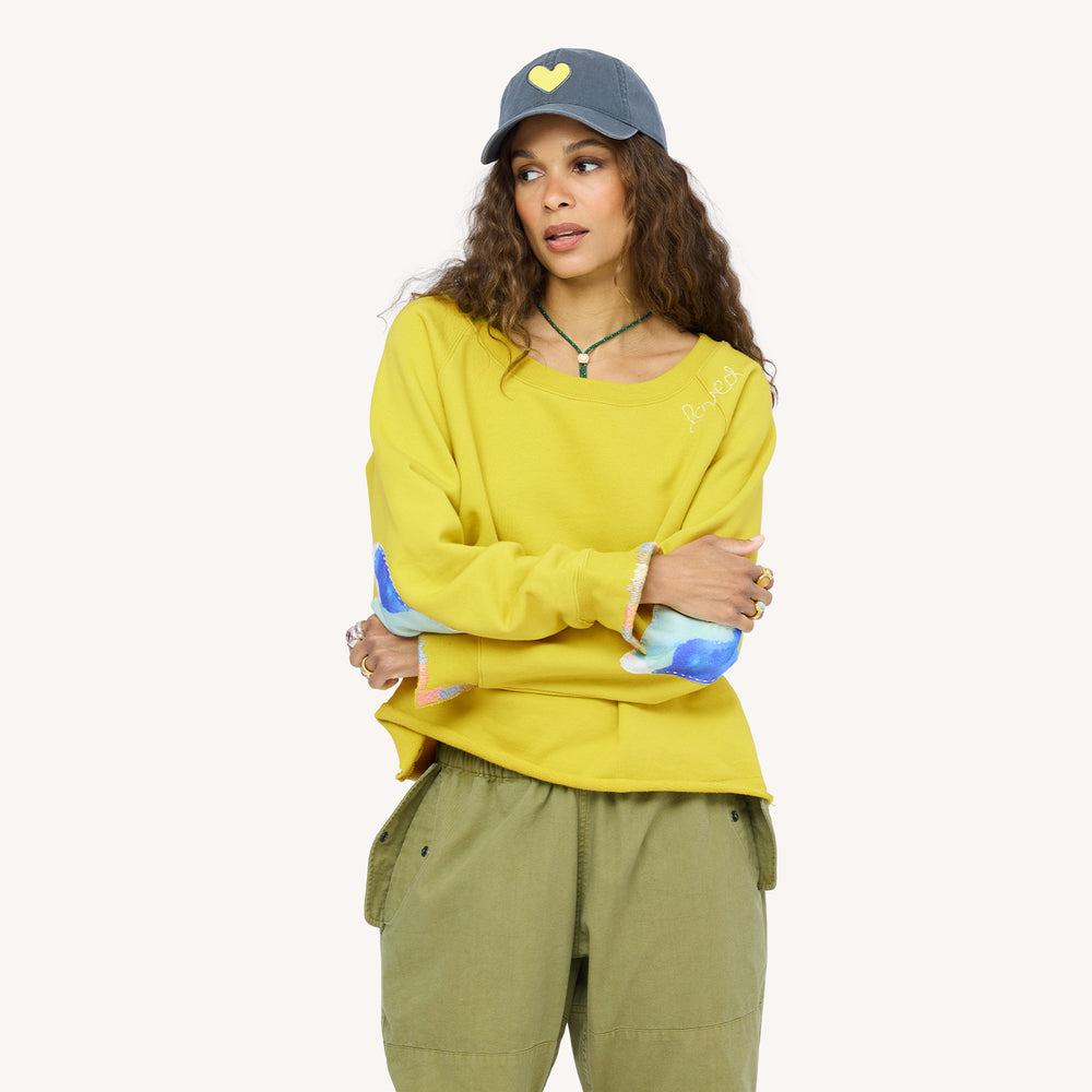 Woman wearing a chartreuse sweatshirt with blue heart elbow patches, a coal heart-patch cap, and olive pants.