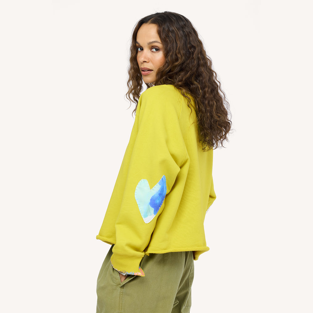 Side back view of a woman wearing a chartreuse sweatshirt with a blue heart elbow patch with paint splats and olive pants.