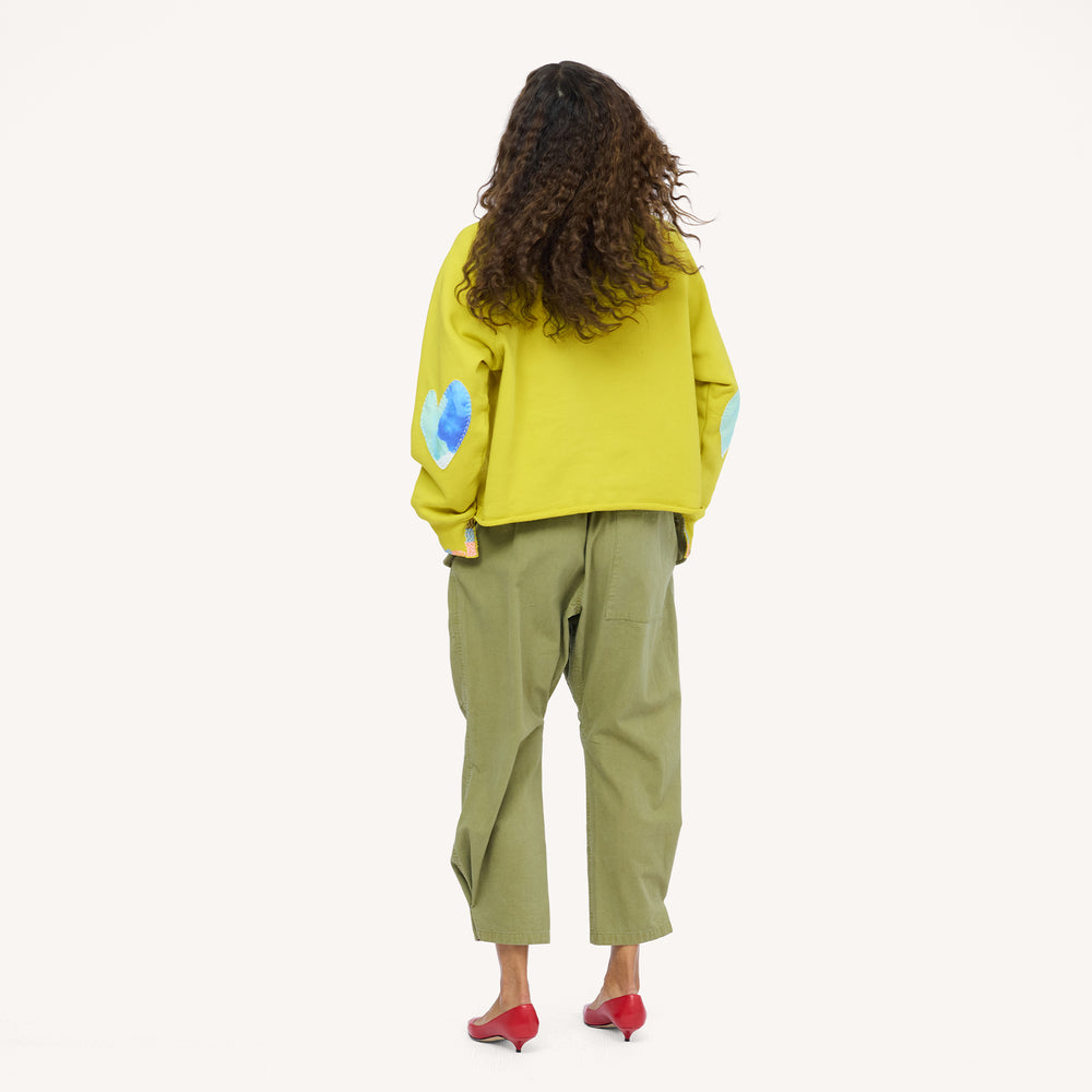 Back view of a woman wearing a chartreuse sweatshirt with blue heart elbow patches, olive pants, and red heels.
