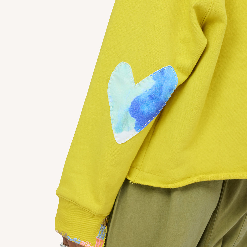 Close up of chartreuse sweatshirt with a blue heart elbow patch, and olive pants.