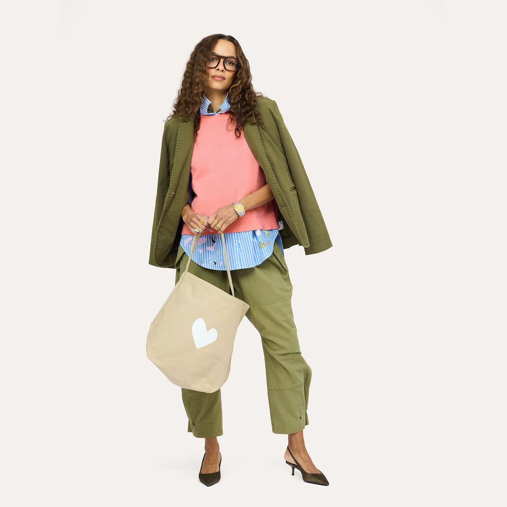 Woman wearing a pink sweater vest layered over a blue striped shirt with an olive blazer and pants, carrying a tan heart tote bag.