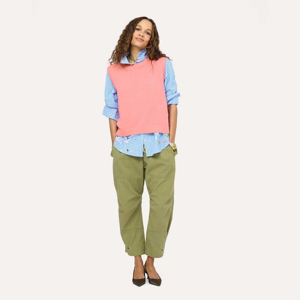 Woman wearing a pink sleeveless sweater vest over blue and white stripe button-down with olive green pants, , and gold watch, smiling against a neutral background.