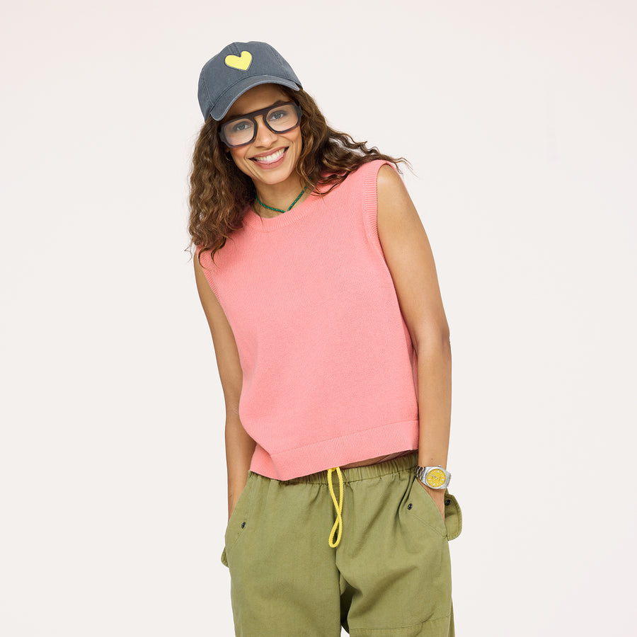 Woman wearing a pink sleeveless sweater vest with olive green pants, a denim heart-patch cap, and gold watch, smiling against a neutral background.