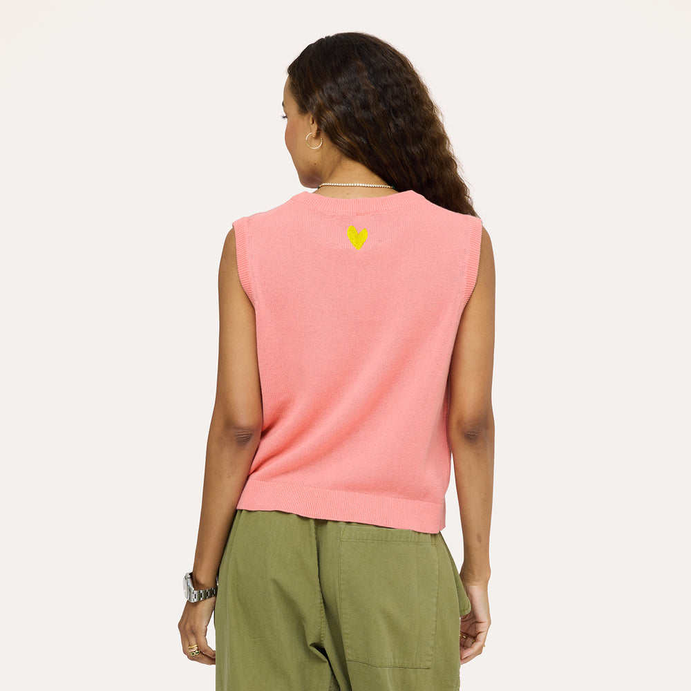 Woman wearing a pink sweater vest from the back with yellow embroidered heart on back and army green pants.