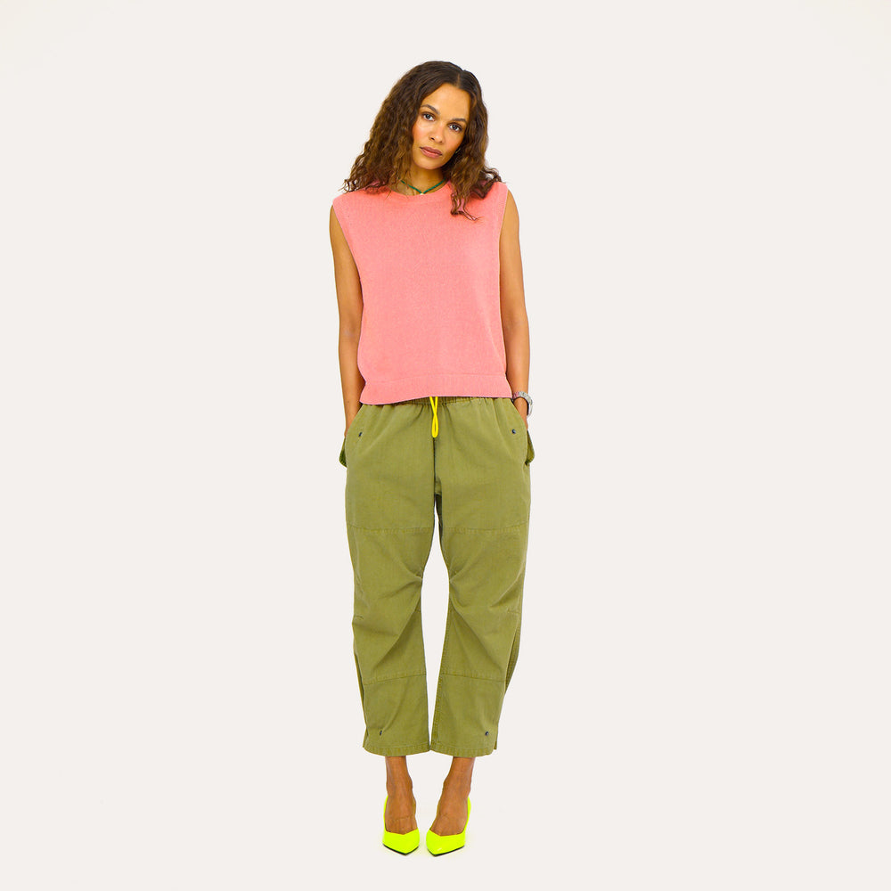 Woman wearing a pink sleeveless sweater vest with olive green pants,  against a neutral background.