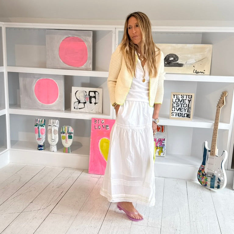 Kerri Rosenthal standing in art gallery wearing a white open-knit crochet cardigan withneon yellow-green trim and  white flowy maxi skirt and white henley tank.