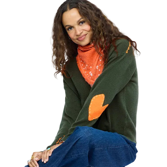 Woman wearing green featherweight cashmere sweater with orange heart elbow patch, styled with an orange bandana. 