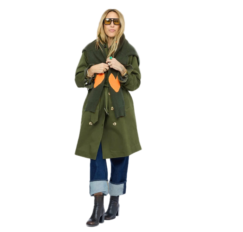 Woman layering the green cashmere sweater with orange heart elbow patches under an olive trench coat, styled with cuffed jeans and ankle boots