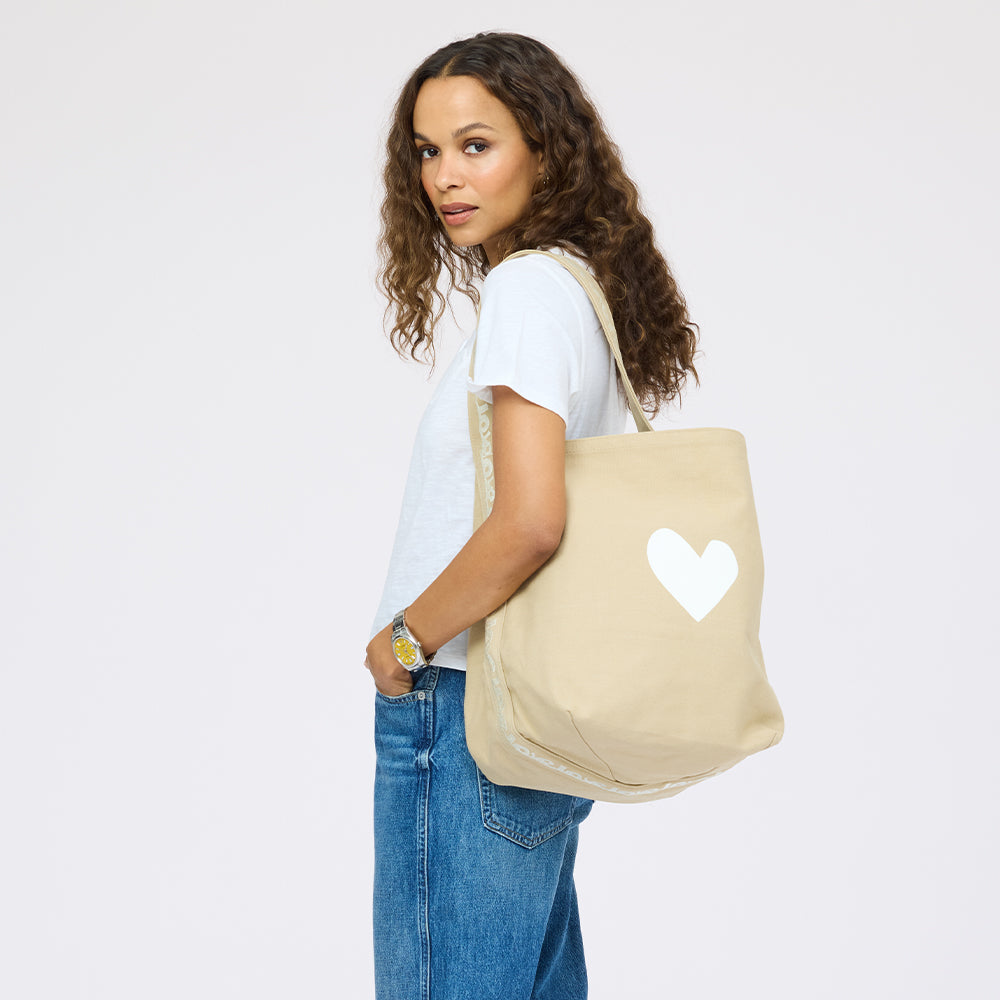 Woman in white tee and denim carrying beige hobo bag with white heart on center.