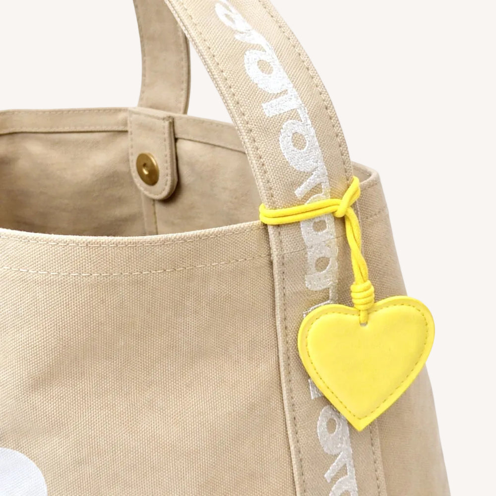 Close-up of a tan canvas tote bag showing the branded strap detail and yellow heart keychain charm.