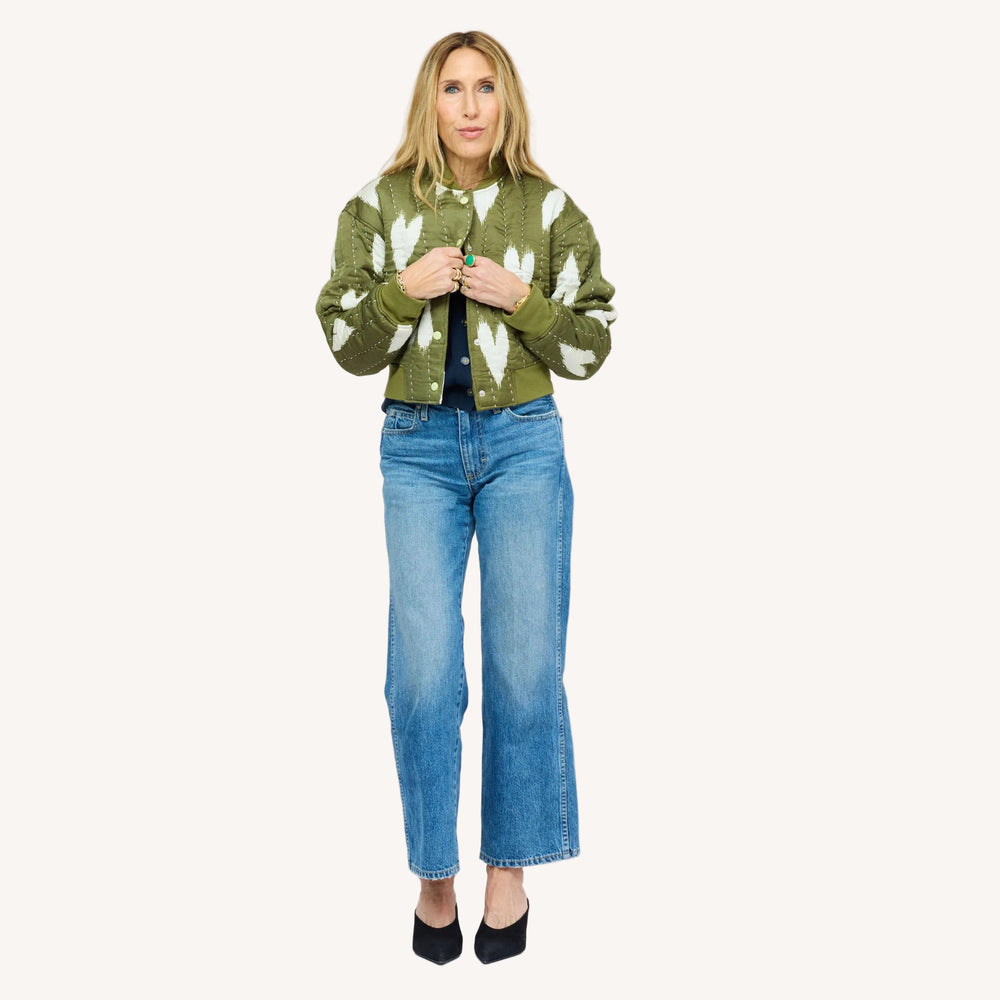 Woman wearing an olive green quilted bomber jacket with heart print, jeans, and black heels.