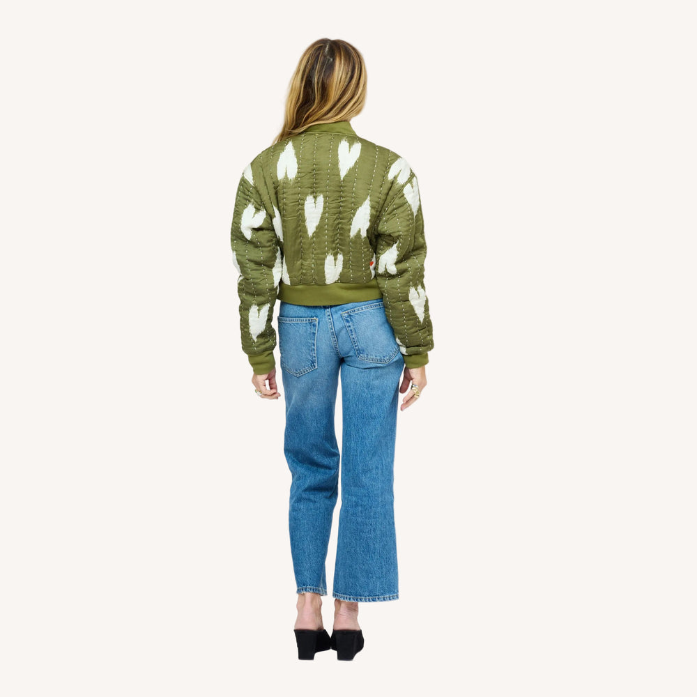 Woman wearing an olive green quilted bomber jacket with heart print, jeans, and black heels. Shop jackets and blazers at Kerri Rosenthal.