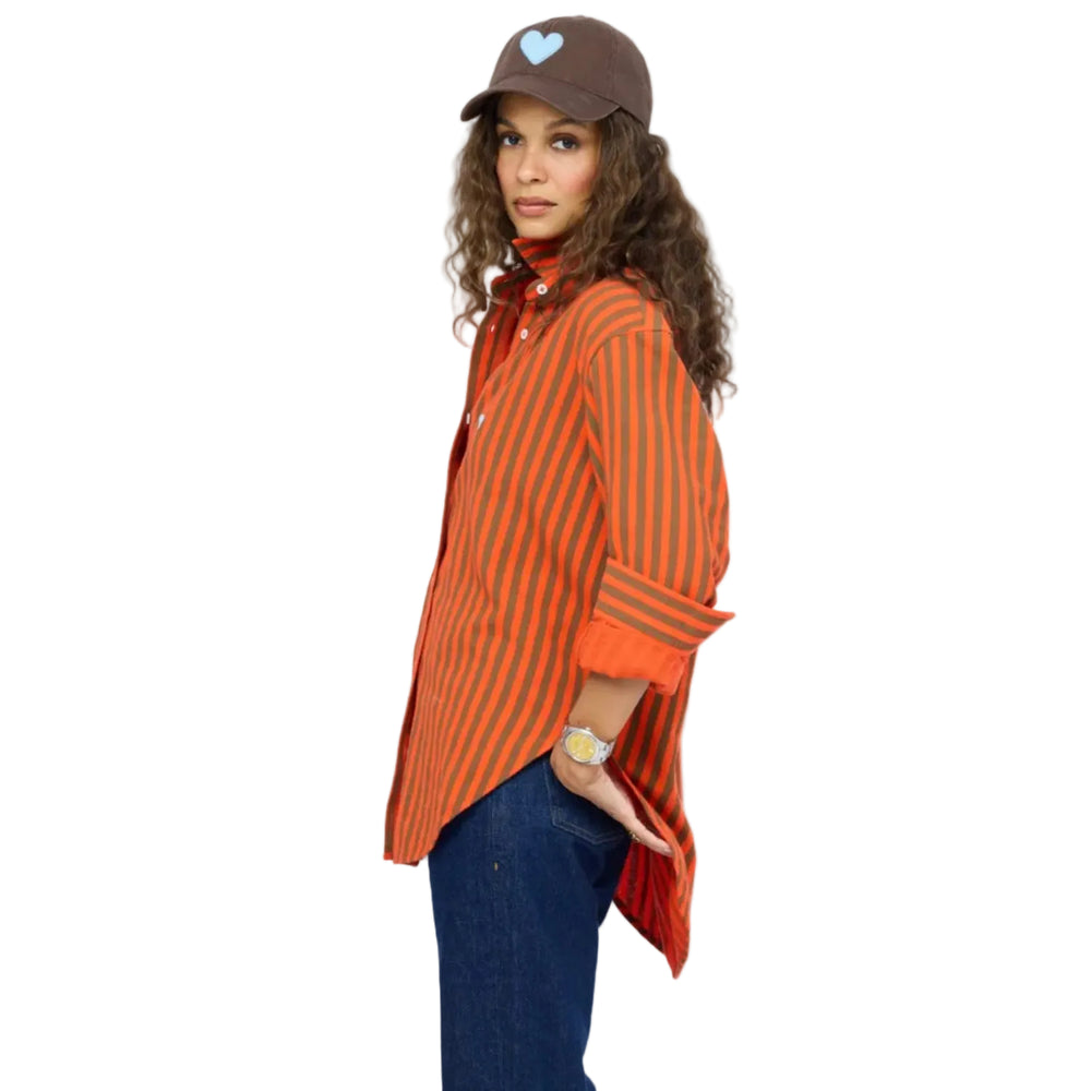 Side view of a woman wearing a Kerri Rosenthal orange and brown striped button-down shirt, dark denim jeans, a brown heart-graphic cap, and a wristwatch.