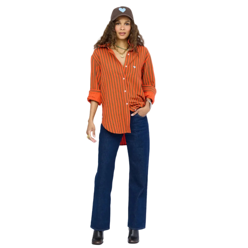 Woman wearing a Kerri Rosenthal orange and brown striped button-down shirt with a small heart embroidery, paired with dark wide-leg jeans, black ankle boots, and a brown heart-graphic cap