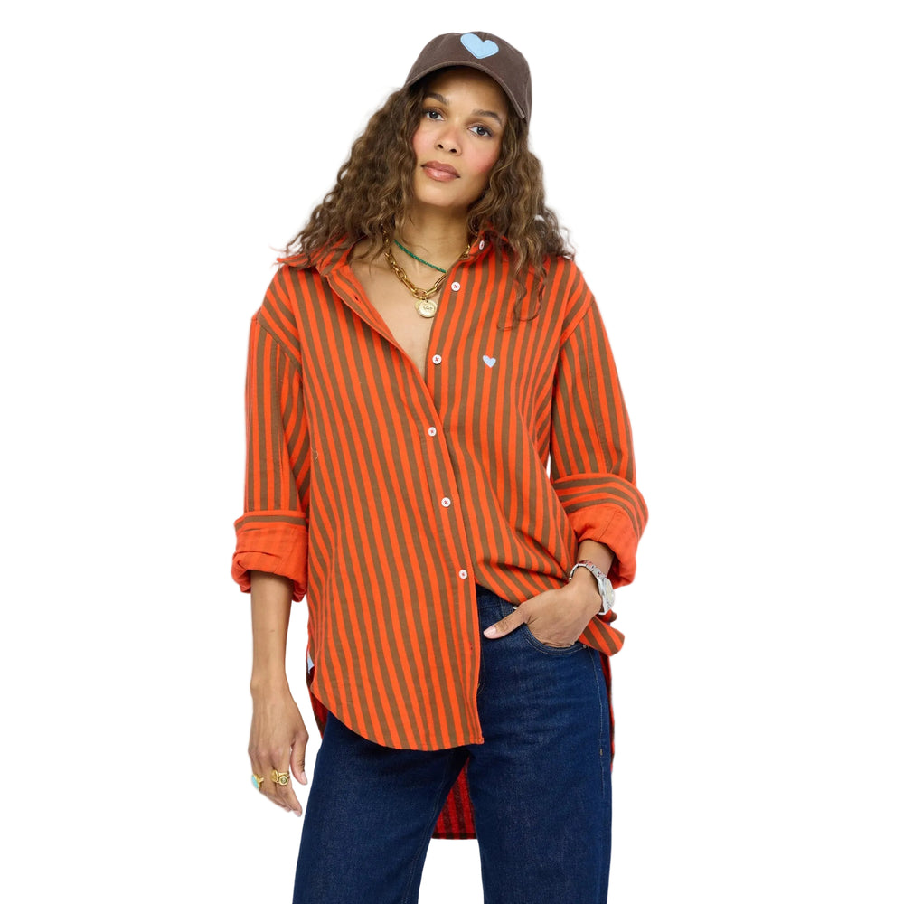 Close-up of a woman wearing a Kerri Rosenthal orange and brown striped button-down shirt with a small heart embroidery, layered gold necklaces, dark denim jeans, a brown heart-graphic cap, and layered rings.