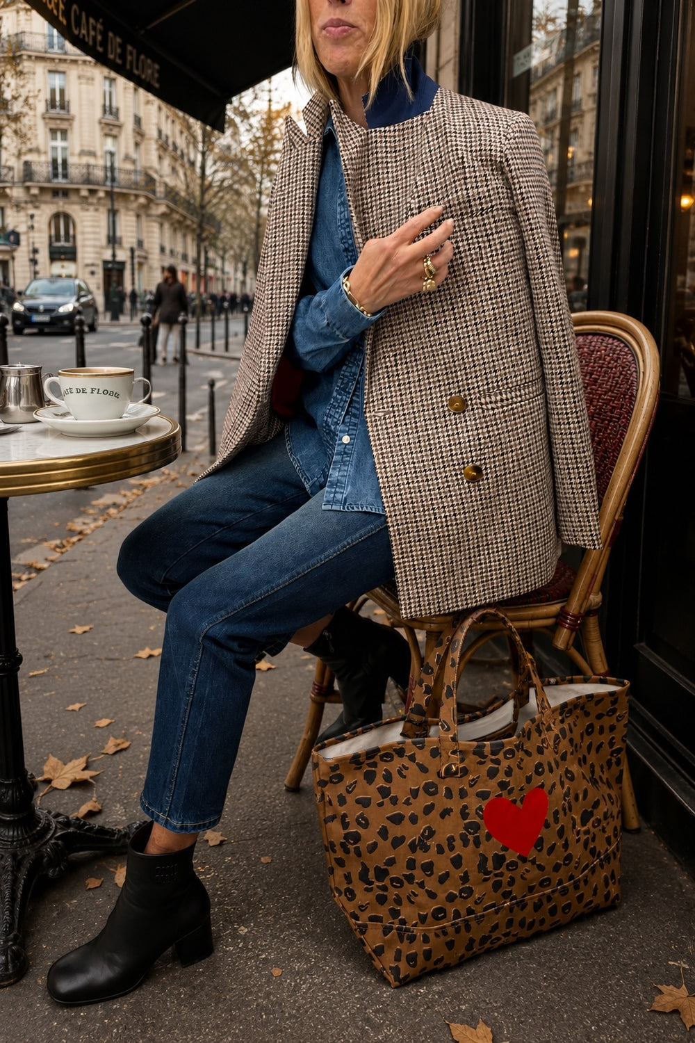 A woman seated on a cafe chair wearing a beige and brown houndstooth blazer, blue denim shirt and jeans, black ankle boots, with a leopard-print tote bag featuring a red heart on the front.
