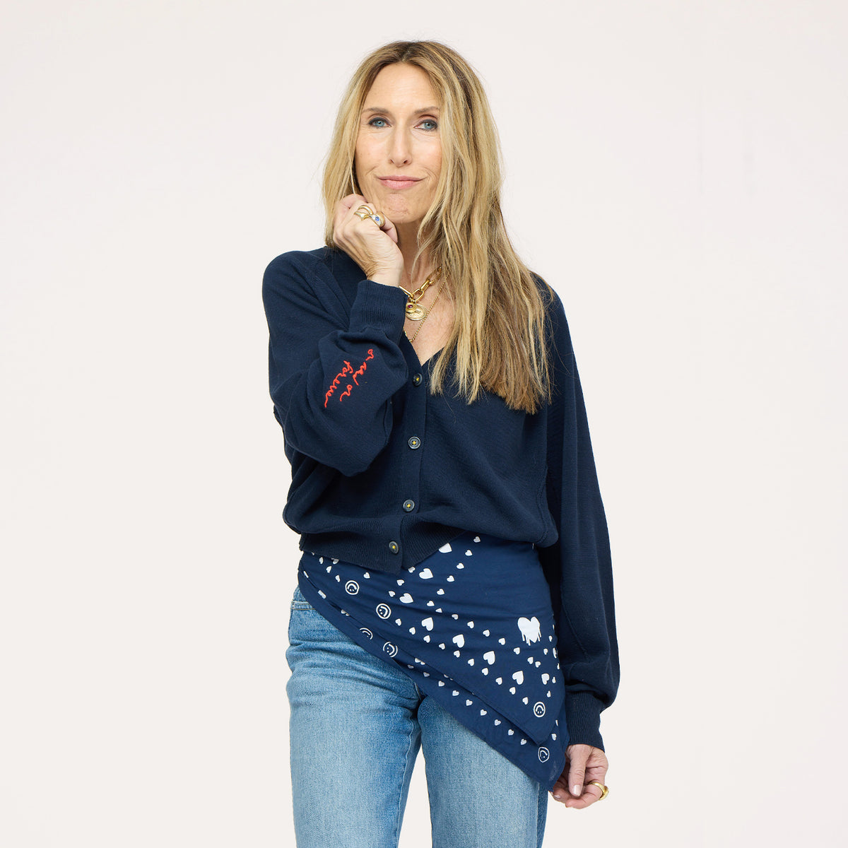 Close-up of woman wearing a Kerri Rosenthal navy v-neck cardigan with red 'now and forever' sleeve embroidery and a navy Kerri Rosenthal heart and smiley print bandana tied at the waist, styled with blue jeans and a gold chain necklace."



