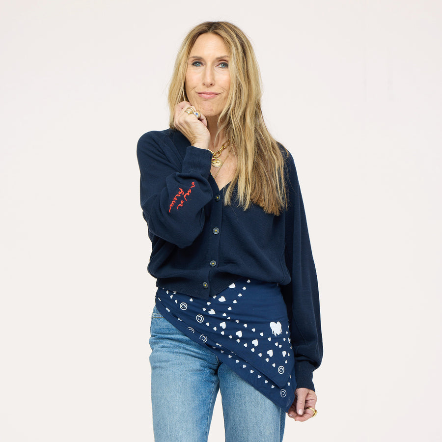 Close-up of woman wearing a Kerri Rosenthal navy v-neck cardigan with red 'now and forever' sleeve embroidery and a navy Kerri Rosenthal heart and smiley print bandana tied at the waist, styled with blue jeans and a gold chain necklace."



