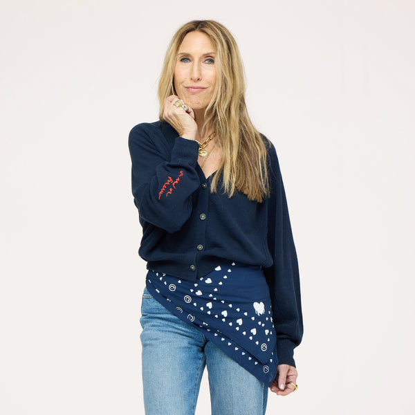 Close-up of woman wearing a Kerri Rosenthal navy v-neck cardigan with red 'now and forever' sleeve embroidery and a navy Kerri Rosenthal heart and smiley print bandana tied at the waist, styled with blue jeans and a gold chain necklace."



