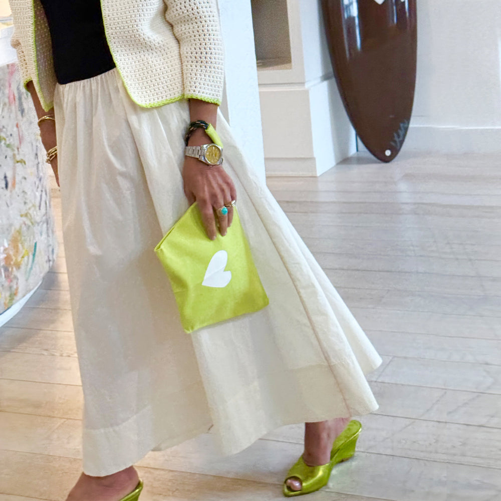 Kerri wearing a cream crochet Frankie cardigan with neon yellow-green trim, a white flowy maxi kr two tone tank dress and lime green wedge sandals, holding a chartreuse heart clutch bag indoors.