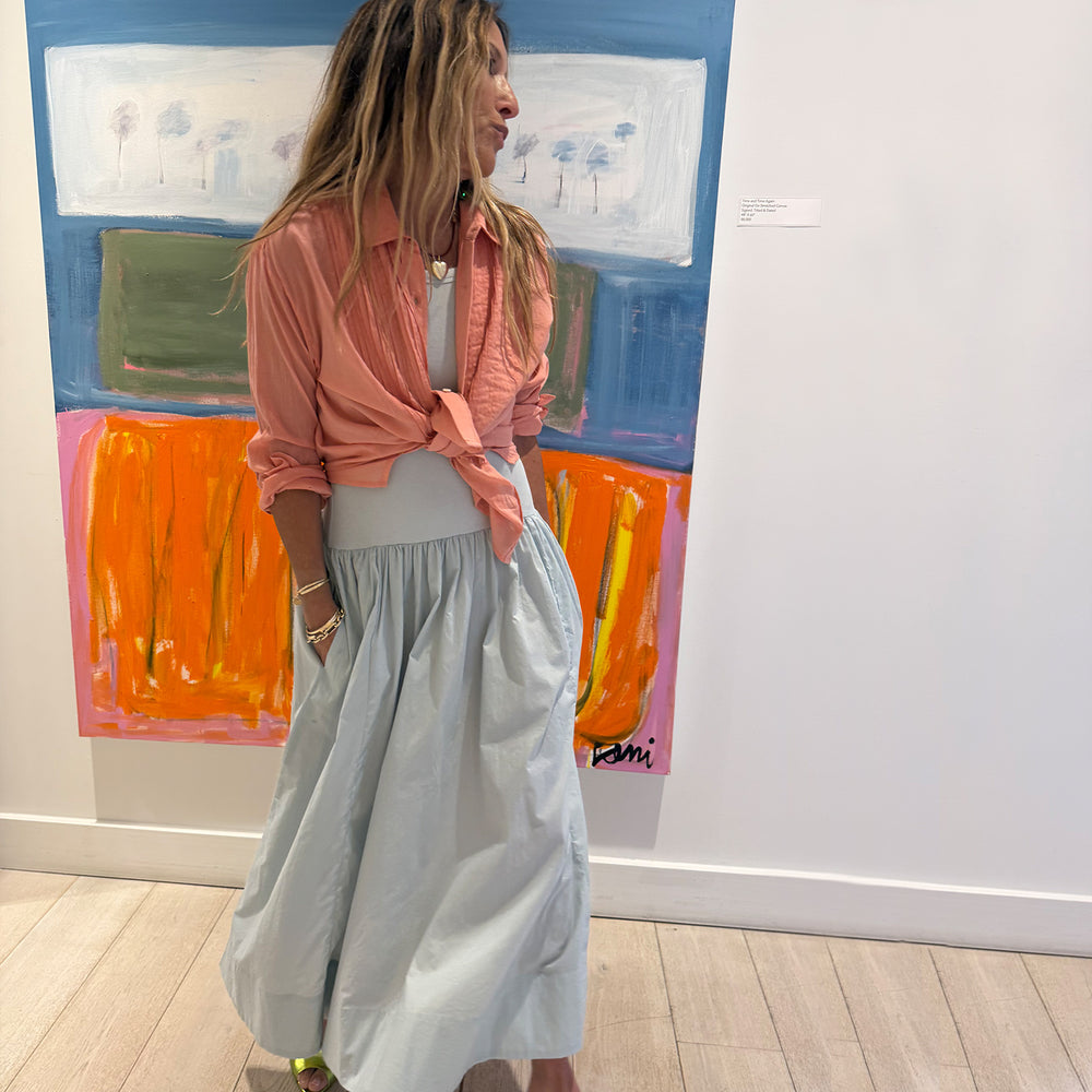 Kerri standing in her Westport gallery wearing a powder blue gathered maxi KR Tank dress and a coral pink tied button-down Tuxedo shirt, styled with gold jewelry and green heeled sandals in front of a colorful abstract painting.